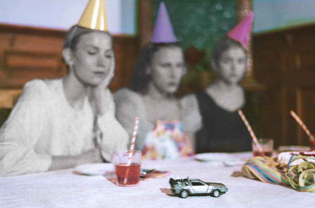 Leikkiauto lastenjuhlien pöydällä, taka-alalla kolme nuorta naista juhlapäähineet päässä