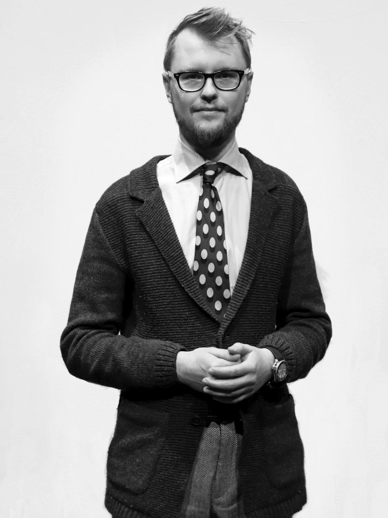 Svart-vit bild på man i glasögon, yllekofta och prickig slips står med händerna knutna framför vit bakgrund.
